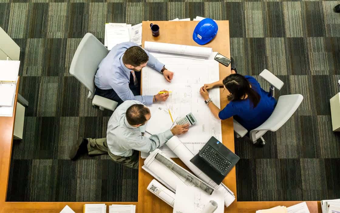 Three colleagues reviewing building plans around a desk with a laptop hard hat and calculator